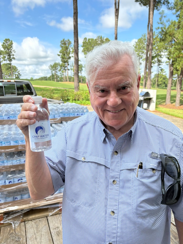 Man holding an 8-ounce bottle of CPAP Water, with a truckload of cases behind him